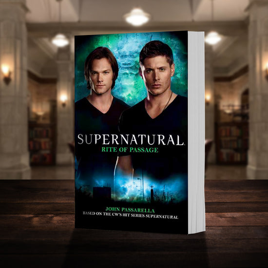 A copy of the book "Supernatural: Rite of Passage" pictured in the "bunker" from Supernatural. The cover shows early-season Sam and Dean Winchester and reads "Supernatural Rite of Passage - Based on the hit CW series SUPERNATURAL by John Passarella”