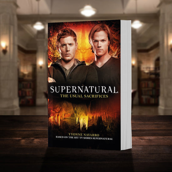 A copy of the book "Supernatural: The Usual Sacrifices" pictured in the "bunker" from Supernatural. The cover shows early-season Sam and Dean Winchester and reads "Supernatural The Usual Sacrifices - Based on the hit CW series SUPERNATURAL by Yvonne Navarro”