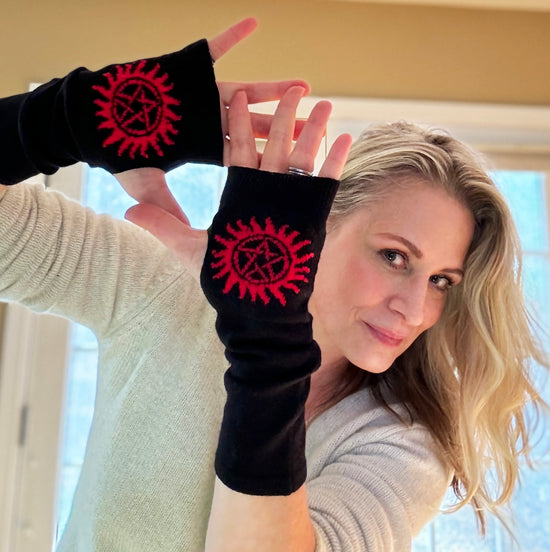 Actress Samantha Smith in a light grey sweater, showing off a pair of fingerless, black gloves with red anti-possession symbols on the palms. The gloves are a few inches longer than wrist-length. 