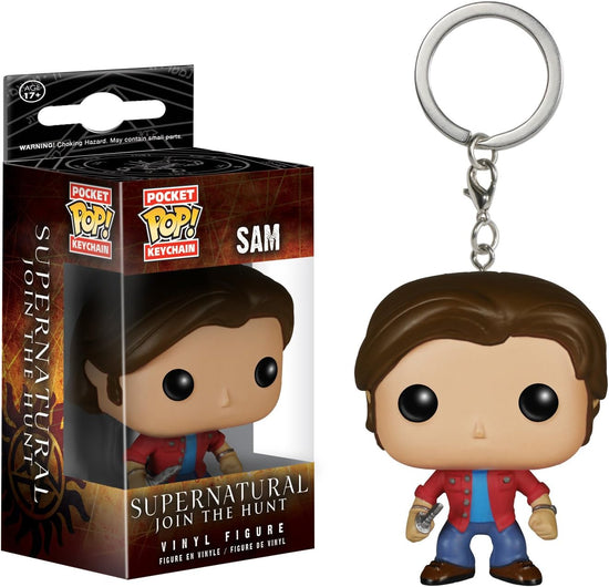 Close up of a keychain in the shape of Sam Winchester, wearing jeans and a red shirt. His features are cartoonish, with an extra large head. Next to the keychain is a brown box containing the same keychain. At the bottom of the box is white text saying "Supernatural, Join the Hunt. Vinyl figure."
