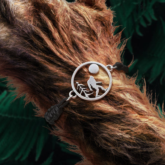 A corded bracelet with a silver, circular charm of Bigfoot walking past a tree under the full moon. The bracelet is attached to a large wrist covered in brown fur