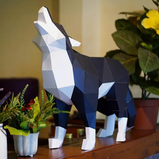 A 3D paper model of a white and black howling wolf. The wolf is standing on a wooden ledge, with potted plants next to it.