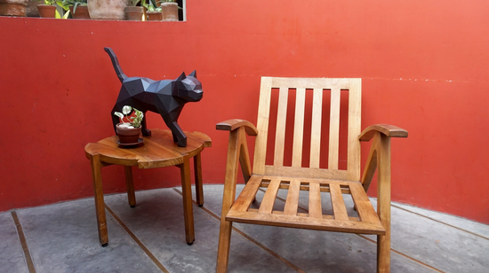A paper model of a black cat is standing on a wooden outdoor table, next to a potted plant. Beside the table is a wooden chair. Behind them is a dark orange wall.