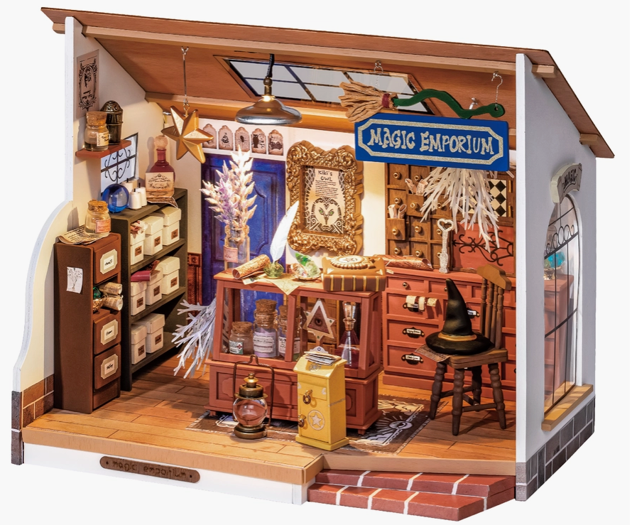 A paper and wood dollhouse showing a magic shop. Magic potions and books are littered about the shop. One wall is covered in cubby holes and drawers. A wooden chair sits in the shop, with a black witch’s hat on the seat.
