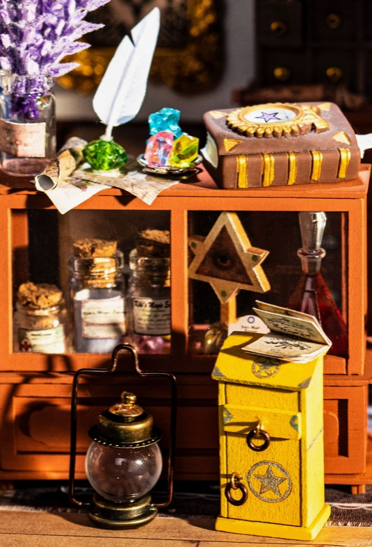 A close up of a cabinet containing tiny glass jars filled with magic ingredients. On top of the cabinet is a brown spell book, with a feather quill and crystals beside it. A miniature gas lantern sits on the floor in front of the cabinet, with a miniature yellow box next to it.