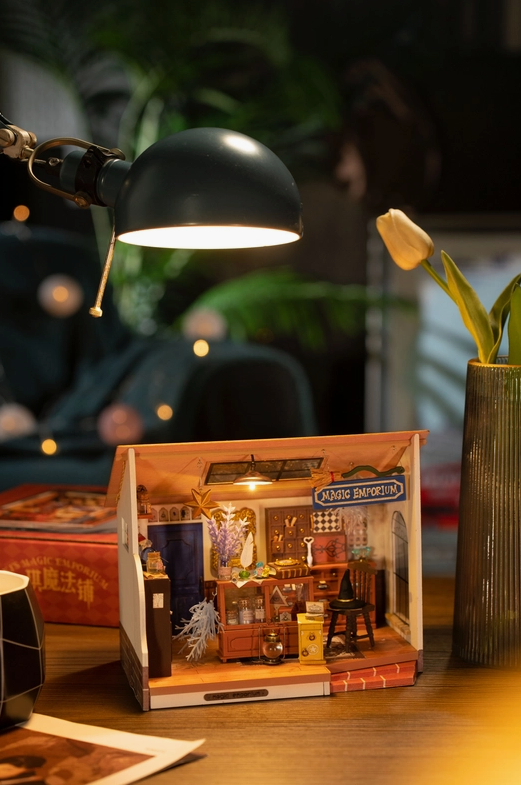An image of the magic emporium dollhouse sitting on a wooden desktop. A desk lamp shines above it.