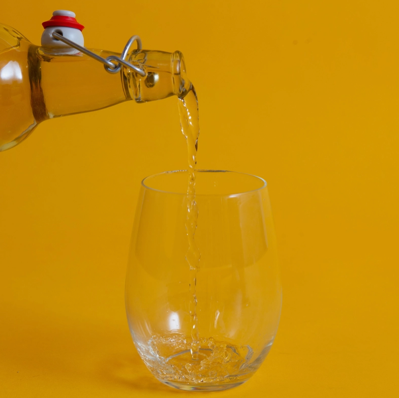 A wine glass on a yellow background. Above the glass is an open bottle, with clear liquid pouring out of it into the glass.