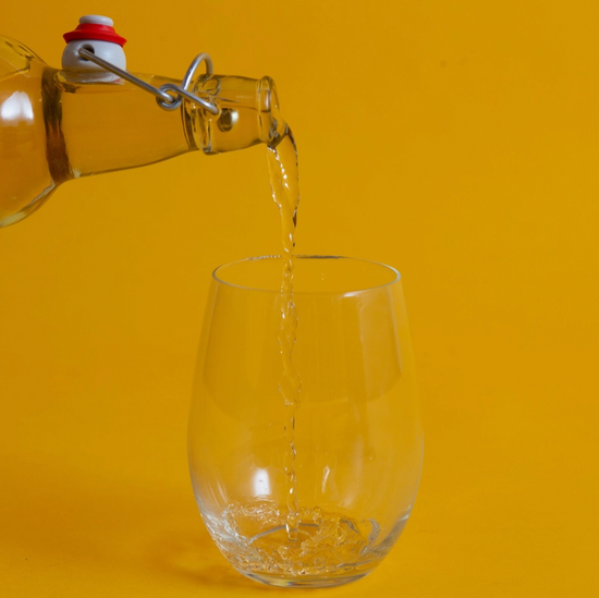 A wine glass on a yellow background. Above the glass is an open bottle, with clear liquid pouring out of it into the glass.