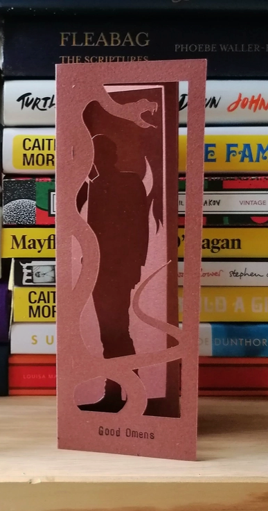 An image of a red bookmark standing in front of a pile of books. The bookmark is cut to depict Crowley, a demon from the series "Good Omens."