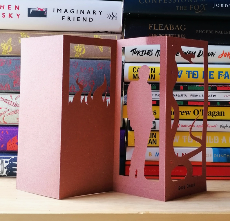 An image of a red bookmark standing in front of a pile of books. The bookmark is cut to depict Crowley, a demon from the series "Good Omens."