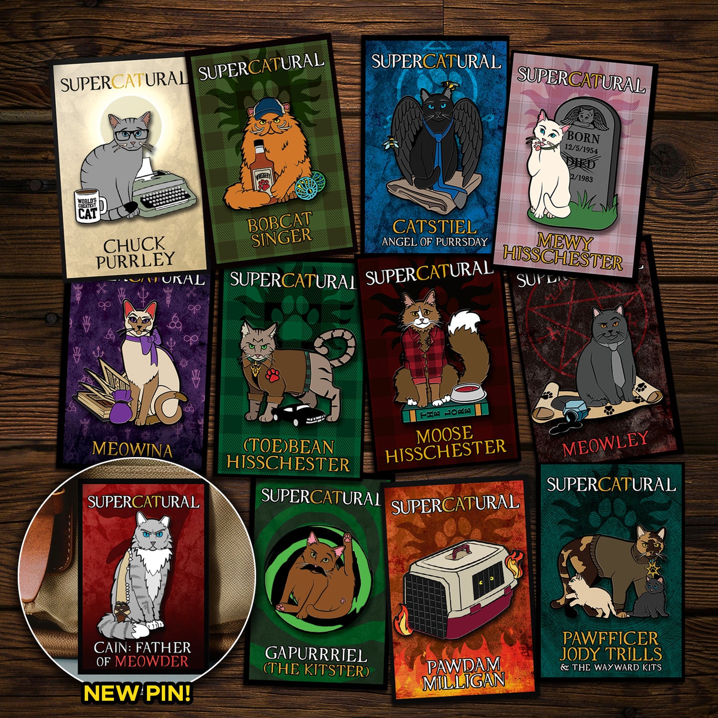 A collection of enamel pins on a wooden table. Each pin depicts a character from the TV series "Supernatural" reimagined as cats. An inset in the bottom left corner reads "NEW PIN!" and has an image of Cain, the newest pin.