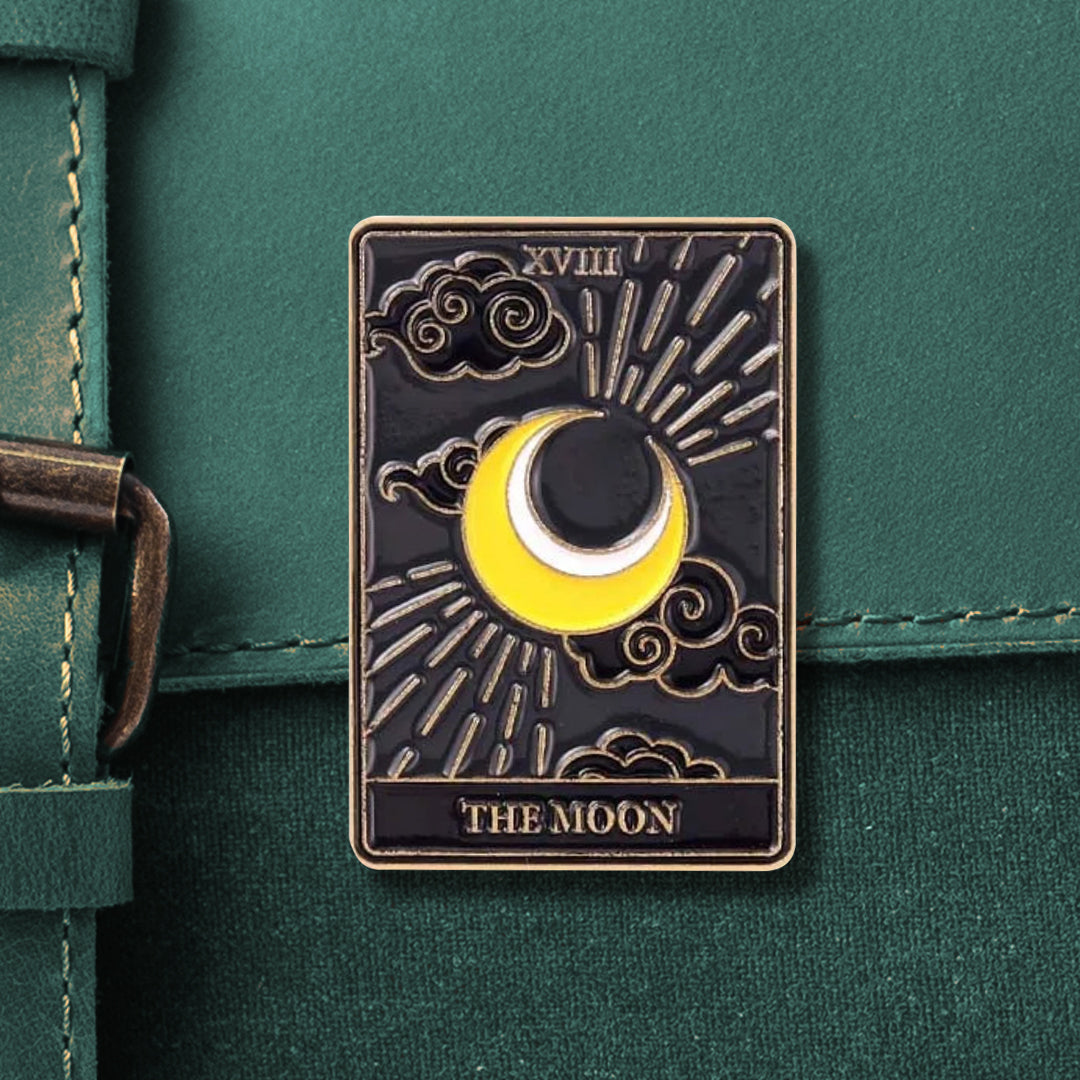 A rectangular tarot pin that reads "XVIII" on the top and "THE MOON" on the bottom, in gold. The pin depicts a yellow-and-white crescent moon surrounded by black-and-gold. swirling clouds, and gold radiance lines. 
