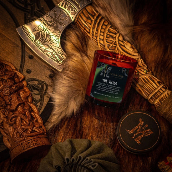 A glass candle lying on a wood table, with a green label. White text on the label says "The viking," with a desciption underneath. Next to the candle is a green lid. Around the candle are an ornate Viking axe, a white animal fur, a wooden shield, and a wooden arm brace.