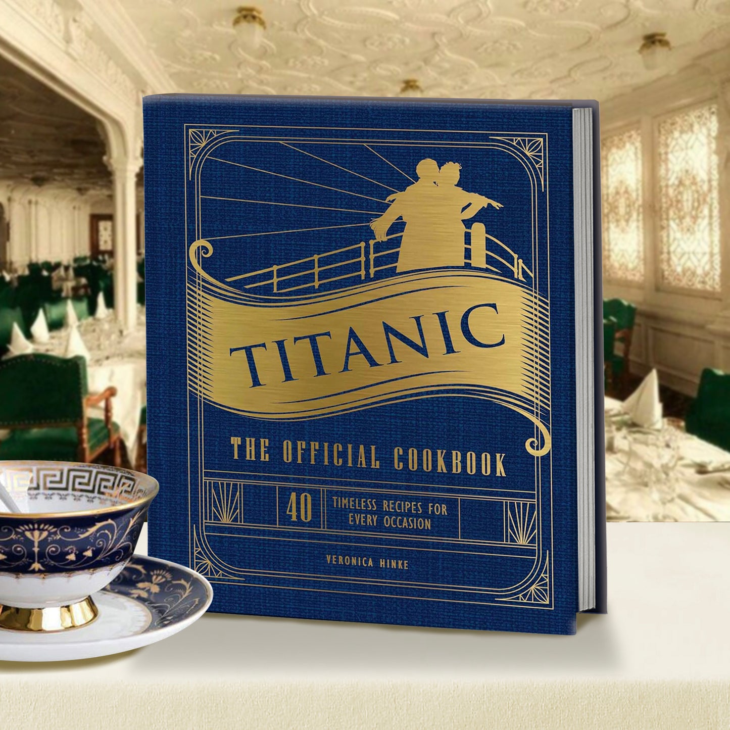 A blue book sitting on a fine dining table. The cover has gold text saying "titanic: the official cookbook." A gold-colored silhouette of Jack and Rose from the movie "titanic" is at the top. There is a blue and white saucer and tea cup sitting to the left of the cookbook.