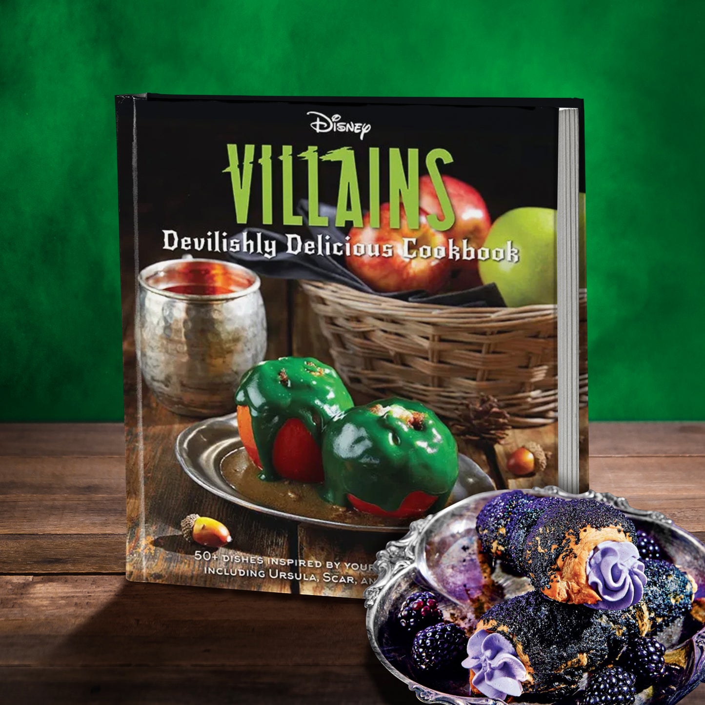 A book on a wood table, in front of a green background. On the book cover is white and green text saying "disney villains, devilishly delocious cookbook." Below the text are apples covered in green ooze, on a metal plate. Behind them is a basket filled with fresh apples. Next to the basket is a metal mug. At the bottom is white text saying "50 plus dishes inspired by your favorite villains, including ursuala, scar, and cruella de vil." Beside the book is a shell filled with flowers and dark berries.