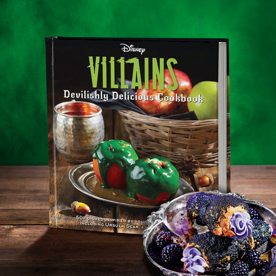 A book on a wood table, in front of a green background. On the book cover is white and green text saying "disney villains, devilishly delocious cookbook." Below the text are apples covered in green ooze, on a metal plate. Behind them is a basket filled with fresh apples. Next to the basket is a metal mug. At the bottom is white text saying "50 plus dishes inspired by your favorite villains, including ursuala, scar, and cruella de vil." Beside the book is a shell filled with flowers and dark berries.