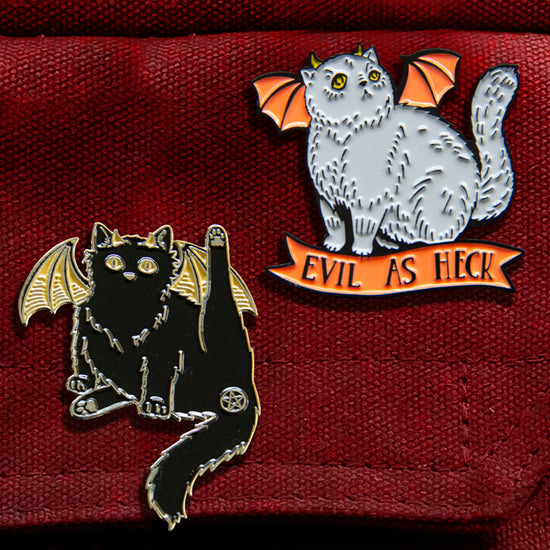Two enamel pins attached to a red canvas bag. One pin is in the shape of a black cat, with yellow bat-like wings. One of the cat’s hind legs is lifted, and the cat’s butthole is covered by a pentagram. The other pin is in the shape of a grey cat with orange bat wings and yellow horns. Under the cat is an orange ribbon with black text saying "Evil as heck."