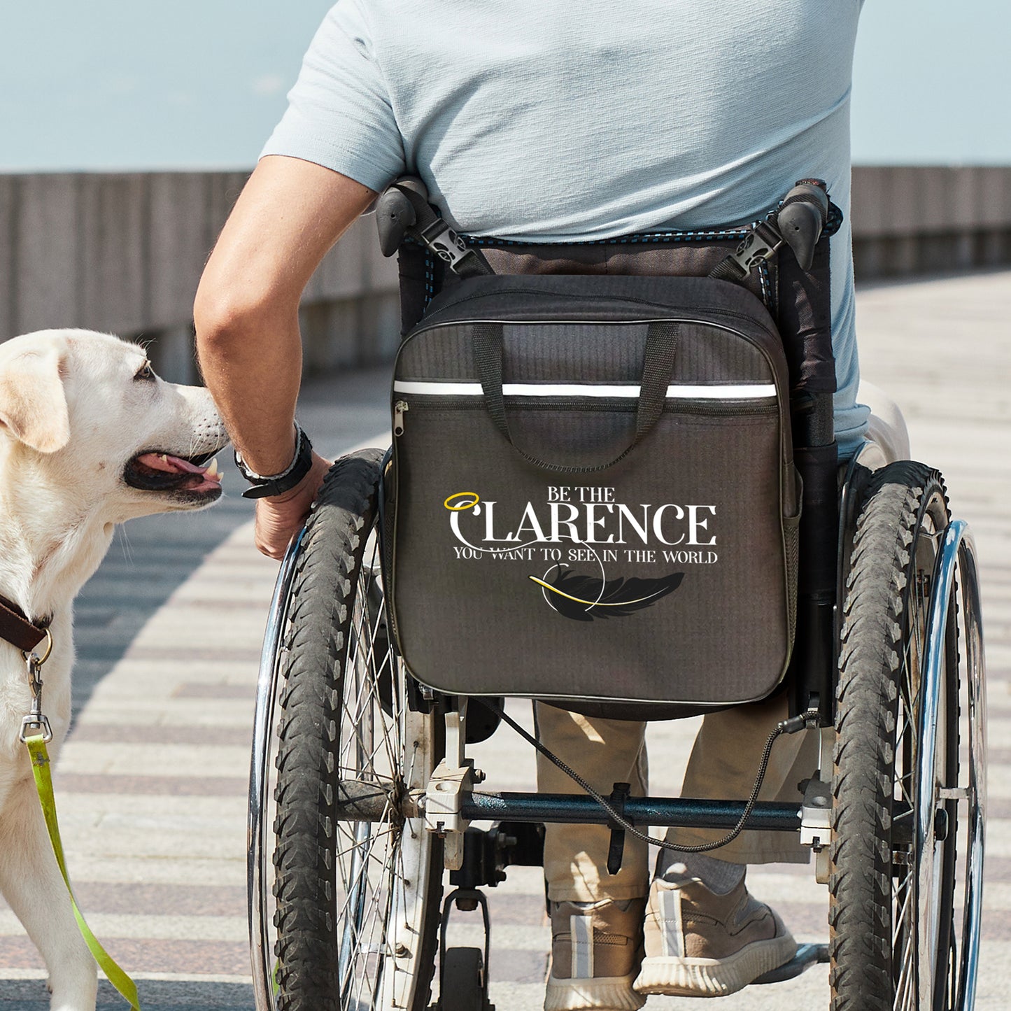 An image of a model in a wheelchair. On the back of the wheelchair is a black messenger-style bag. On the outward-facing side of the bag is the phrase "Be the Clarence you want to see in the world", with a halo over the 'C' and a black feather below the text.