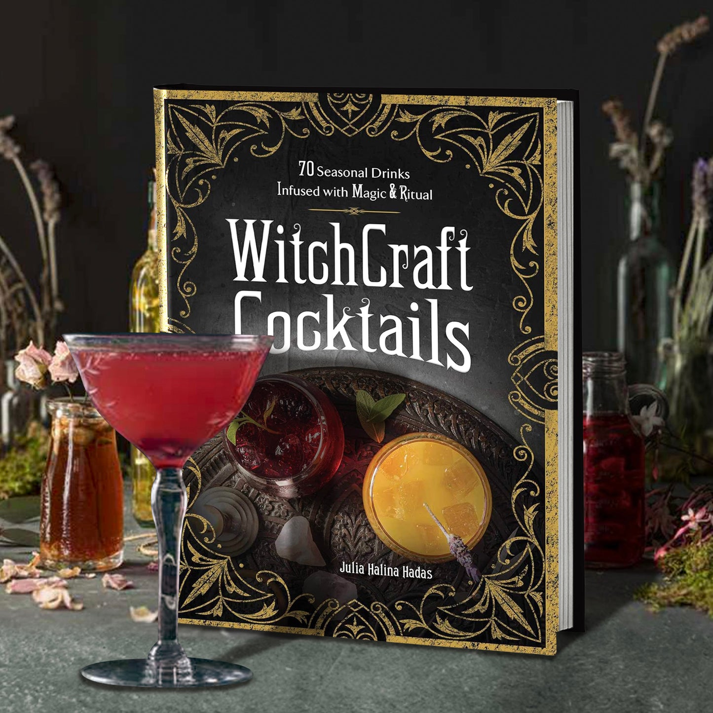 A hardcover book standing on a slate table. The cover has a pair of cocktails on a dark plate, surrounded by an ornate rustic old border. White text says "Witchcraft cocktails." In front of the book is a glass goblet containing a red beverage. Next to and behind the book are glass vials and bottle with various ingredients.