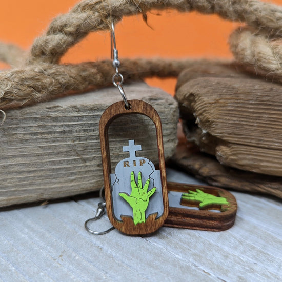 A pair of oval-shaped wooden earrings on a table, leaning aginast driftwood. The center of the earrings are open, with a green zombie hand reaching upward in front of a tombstone.
