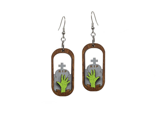 A pair of oval-shaped wooden earrings on a white background. The center of the earrings are open, with a green zombie hand reaching upward in front of a tombstone.