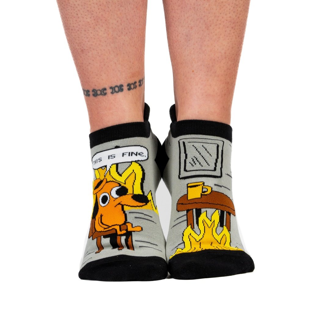 A model's feet wearing a pair of grey and back socks. One sock has a cartoon of a smiling brown dog wearing a hat, sitting in a chair in front of burning flames. Above him is a caption saying "This is fine." The other sock has a brown wooden table with a coffee cup, and a roaring flame underneath it. The cuff and toes of the socks are black.