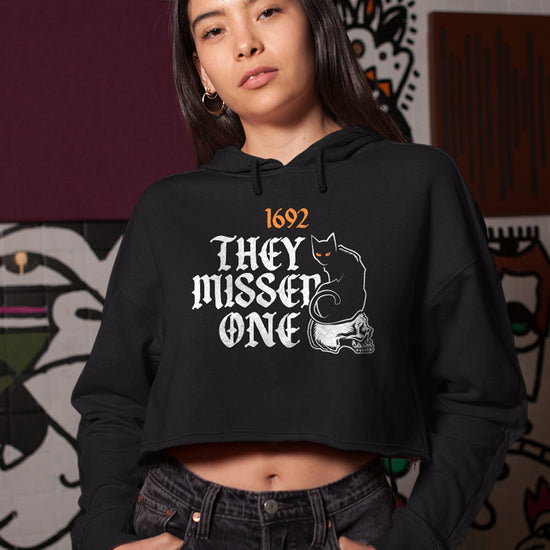A female model wearing a black cropped hoodie. On the hoodie is orange and white text saying "1692: they missed one." Next to the text is an embroidered black cat with orange eyes, sitting on a white skull. Behind the model is a wall painted with graffiti style art.