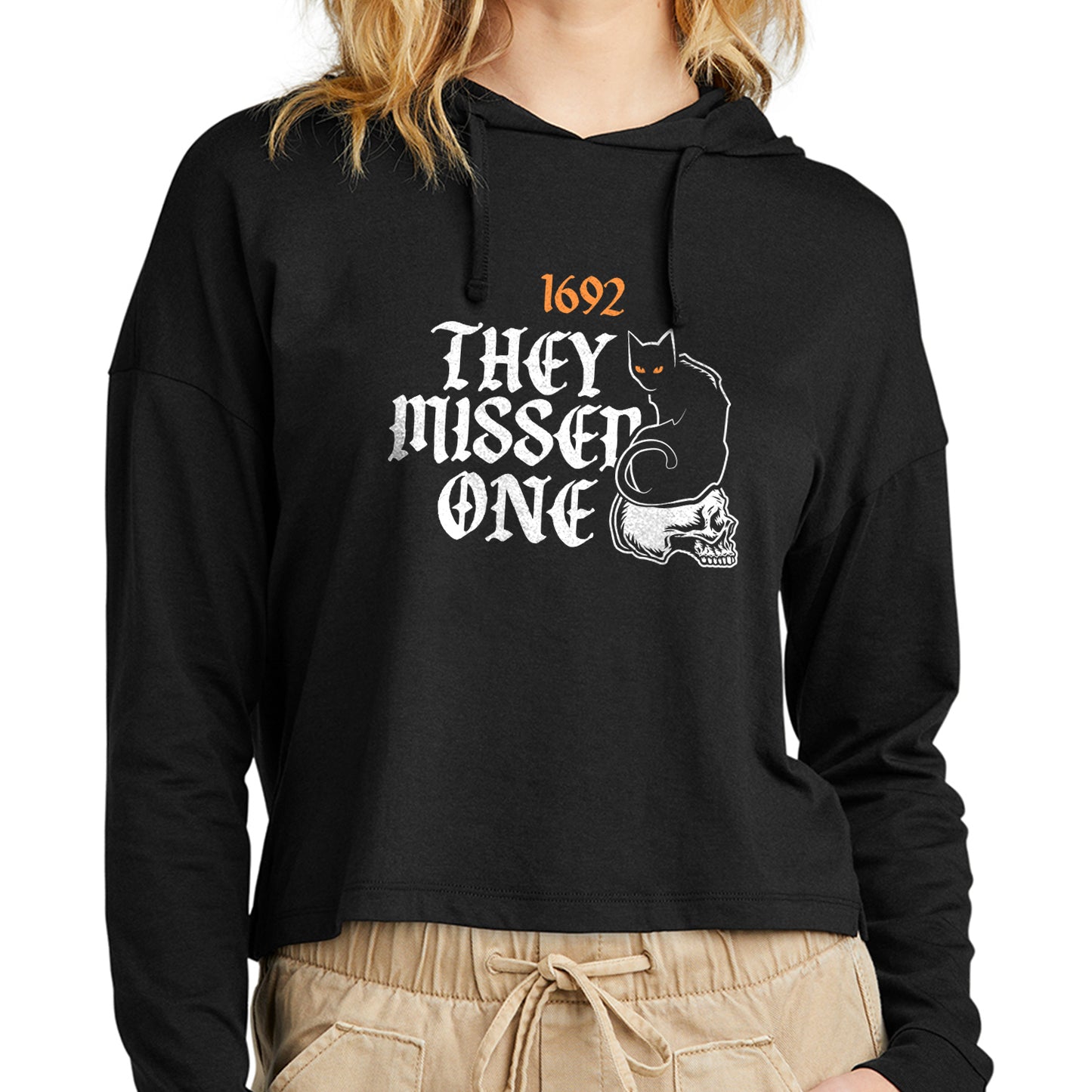 A model wearing a black cropped hoodie, against a white background. On the hoodie is orange and white text saying "1692: they missed one." Next to the text is an embroidered black cat with orange eyes, sitting on a white skull.