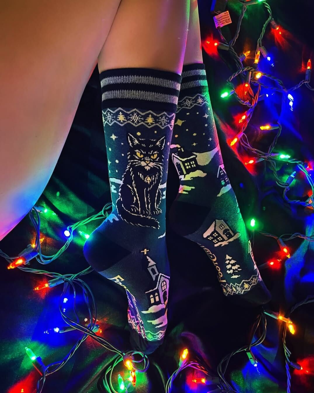 Person wearing festive socks with a cat design, surrounded by colorful Christmas lights.