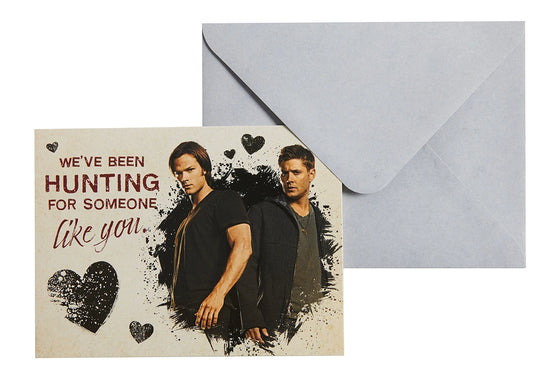 A notecard-style Valentine's card in front of a grey-white envelope. The card shows Sam and Dean on the front and reads "We've been hunting for someone like you"