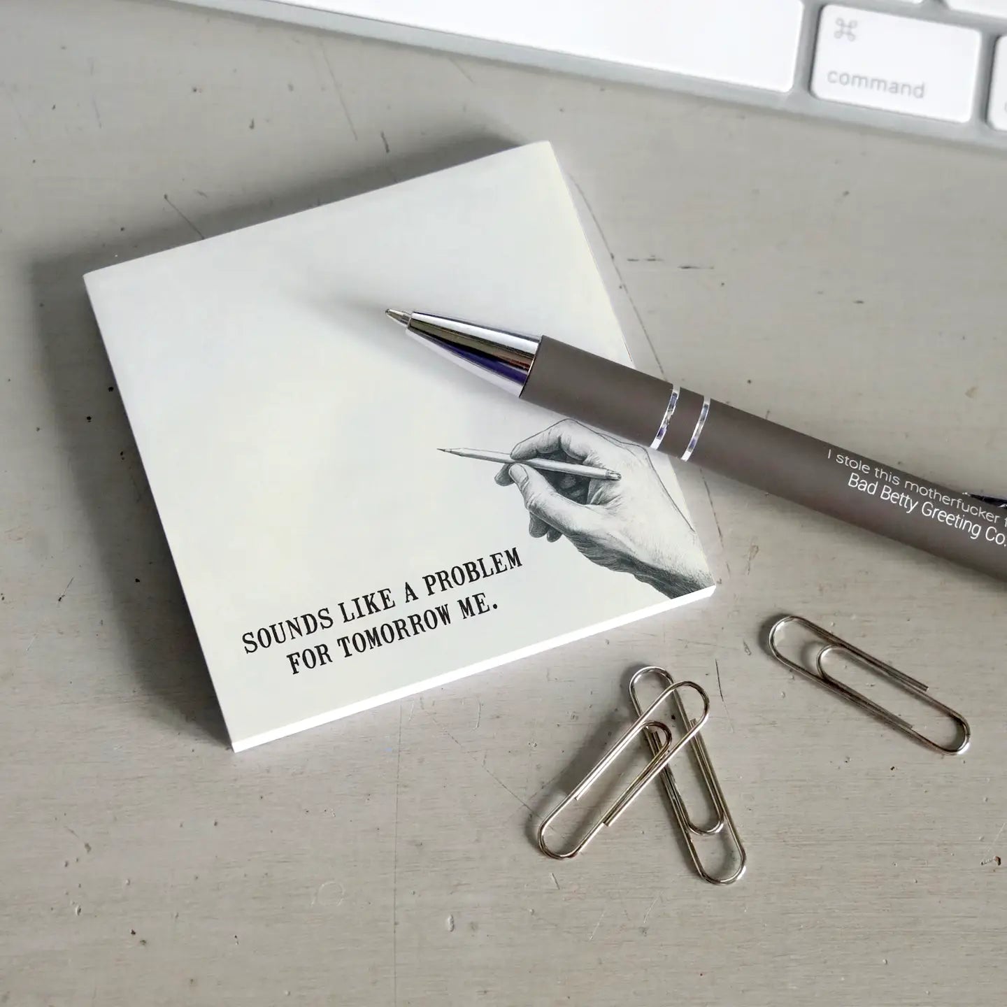 A sticky note with a drawing of a hand holding a pen at the bottom right corner. Text at the bottom says "sounds like a problem for tomorrow me." The note is on a grey desktop. Next to the note is a black pen, a handful of paperclips, and a computer keyboard