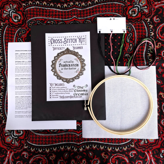 Cross stitch kit with hoop, thread, and pattern on a decorative fabric background. The center has black text saying "actually Frankenstein is the doctor."