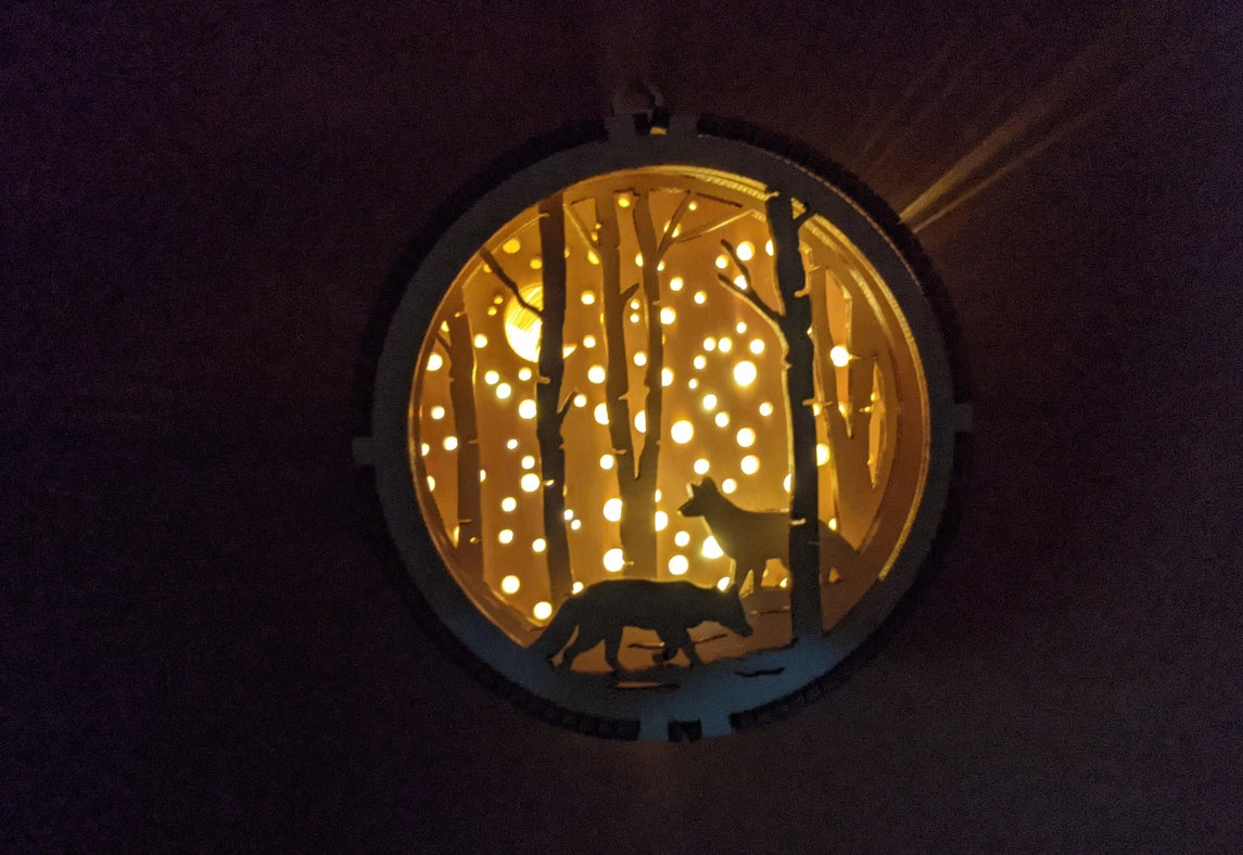 A circular Christmas ornament, hanging against a dark background. The ornament has a 3D depiction of two wolves amongst a forest. The ornament has a light in the background, which peaks between trees and through small holes in the back, depicting snowfall.