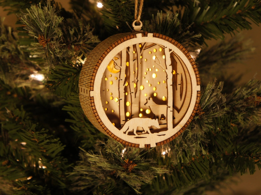A circular Christmas ornament, hanging on a Christmas tree limb. The ornament has a 3D depiction of two wolves amongst a forest. The ornament has a light in the background, which peaks between trees and through small holes in the back, depicting snowfall.