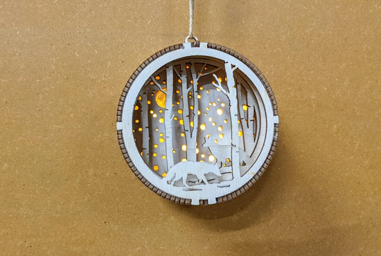 Load image into Gallery viewer, A circular Christmas ornament, hanging against a particle board background. The ornament has a 3D depiction of two wolves amongst a forest. The ornament has a light in the background, which peaks between trees and through small holes in the back, depicting snowfall.
