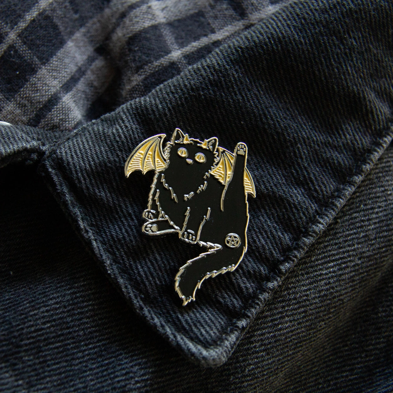 An enamel pin in the shape of a black cat, with yellow bat-like wings, affixed to a dark denim collar. One of the cat’s hind legs is lifted, and the cat’s butthole is covered by a pentagram.