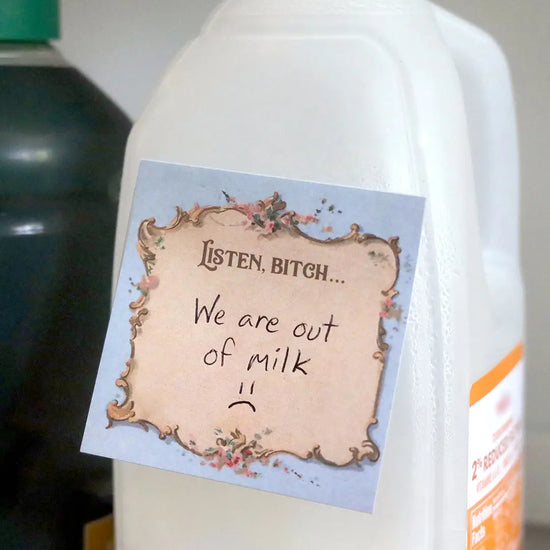 A blue sticky note with a parchment colored center. At the top is flowered filigree, with text saying "Listen, bitch." The note is stuck to a milk carton, with a handwritten note saying "we are out of milk"