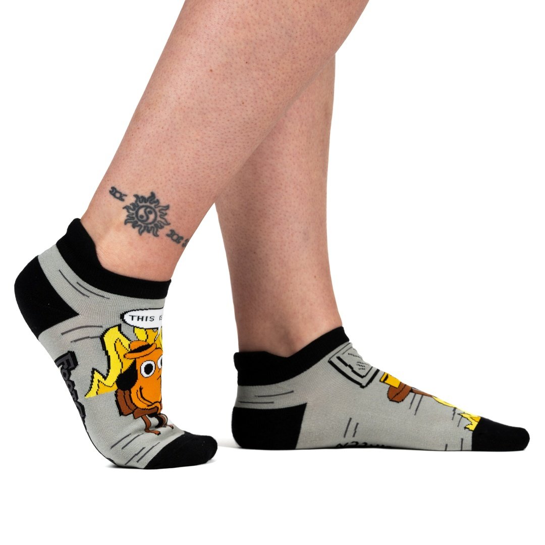 A model's feet wearing a pair of grey and back socks. One sock has a cartoon of a smilingbrown dog wearing a haat, sitting in a chair in front of burning flames. Above him is a caption saying "This is fine." The other sock has a brown wooden table with a coffee cp, and a roaring flame underneath it. The cuff and toes of the socks are black.
