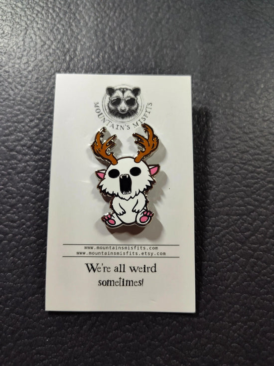 A metal pin depicting a white creature with antlers. The pin is on a white backing card, with black text saying "Mountain's misfits. We're all weird sometimes." The pin is sitting on a black leather surface.