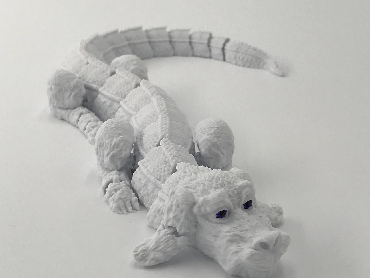 The side view of a light grey, 3D-printed rendition of Falkor the Luckdragon from The NeverEnding Story. It has dark black-purple eyes and printed joints that allow it to act as a fidget toy.