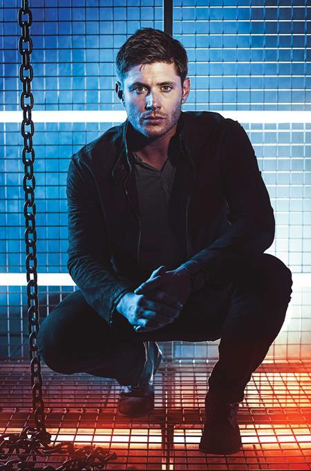 Dean Winchester in a dark jacket sitting on a metal platform with chains, against a tiled wall.