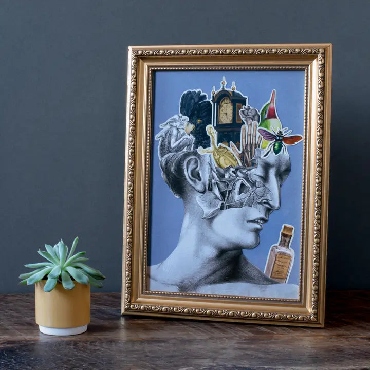 A framed picture on a wooden desk. The picture shows a drawing of a person's head, with various random images affixed to it. In front of the framed picture is a small cactus in a cup.