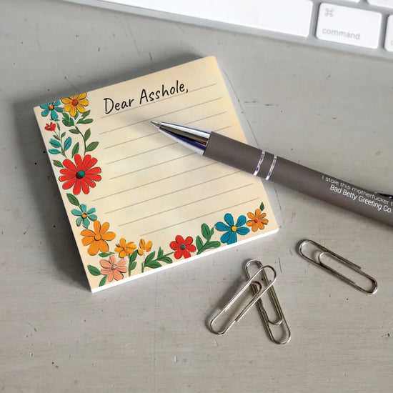 A yellow sticky note with horizontal lines. At the top is black text saying "Dear asshole." At the left and bottom are brightly colored flowers. The note is on a grey desktop. Next to the note is a black pen, a handful of paperclips, and a computer keyboard