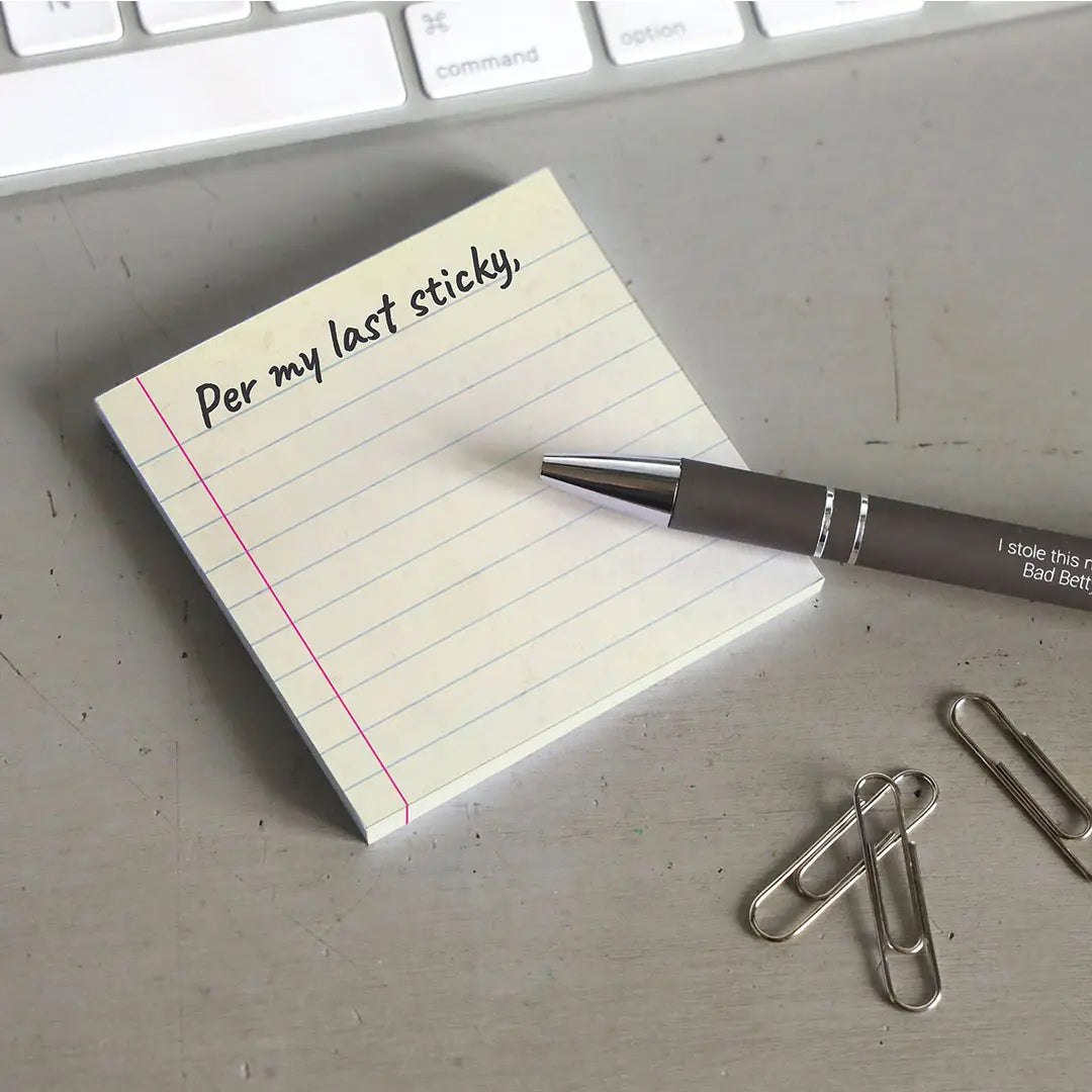 A yellow sticky note with blue horizontal lines. At the top is black text saying "per my last sticky." The note is on a grey desktop. Next to the note is a black pen, a handful of paperclips, and a computer keyboard
