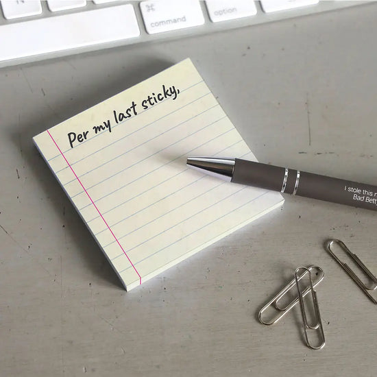 A yellow sticky note with blue horizontal lines. At the top is black text saying "per my last sticky." The note is on a grey desktop. Next to the note is a black pen, a handful of paperclips, and a computer keyboard
