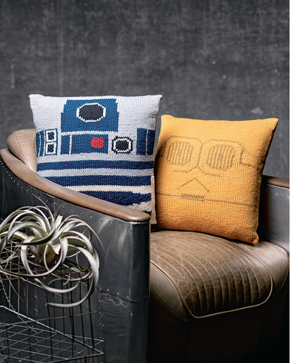 Two knit throw pillows on a leather chair. One pillow is the blue and white face of R2D2, the other is the gold face of C3PO, two droids from the Star Wars movies.