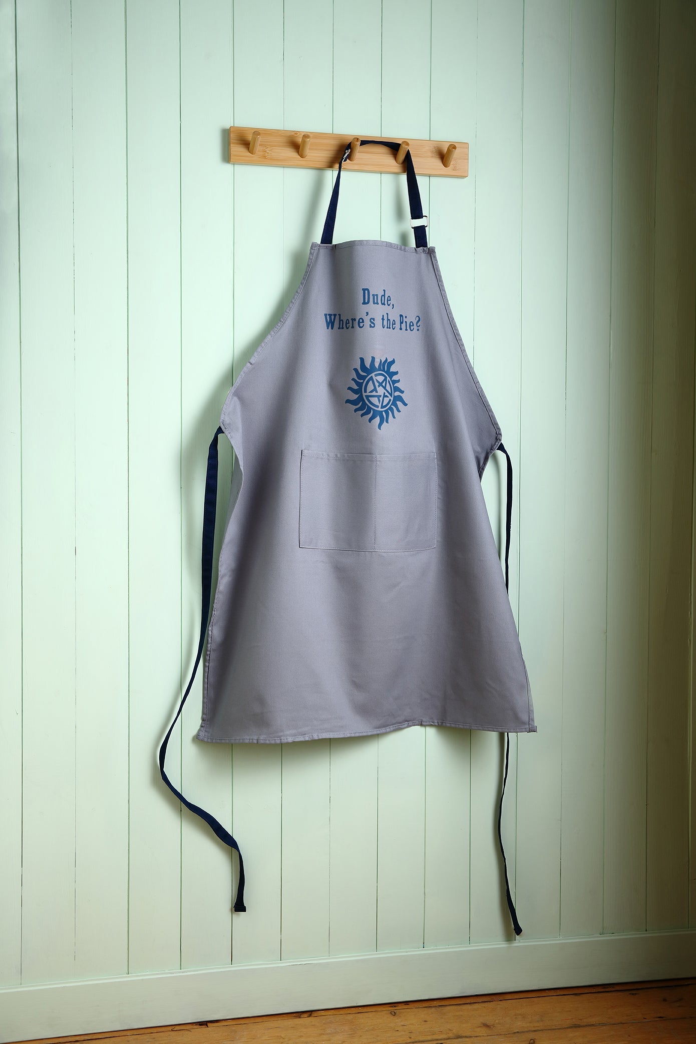 Gray apron with blue design and text hanging on a wooden hook against a light green wall.