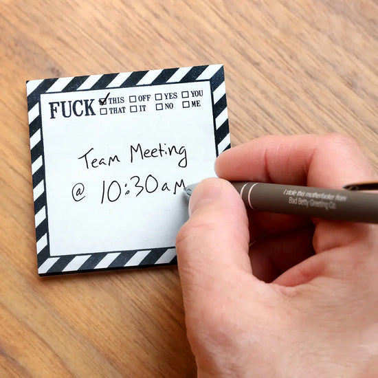 A white sticky note with black diagonal lines around the border. At the top is black text saying "fuck" with various checkable options next to it, including "this, off, you, and no". The note is on a wooden desktop. A hand holding a pen is writing a note on the sticky