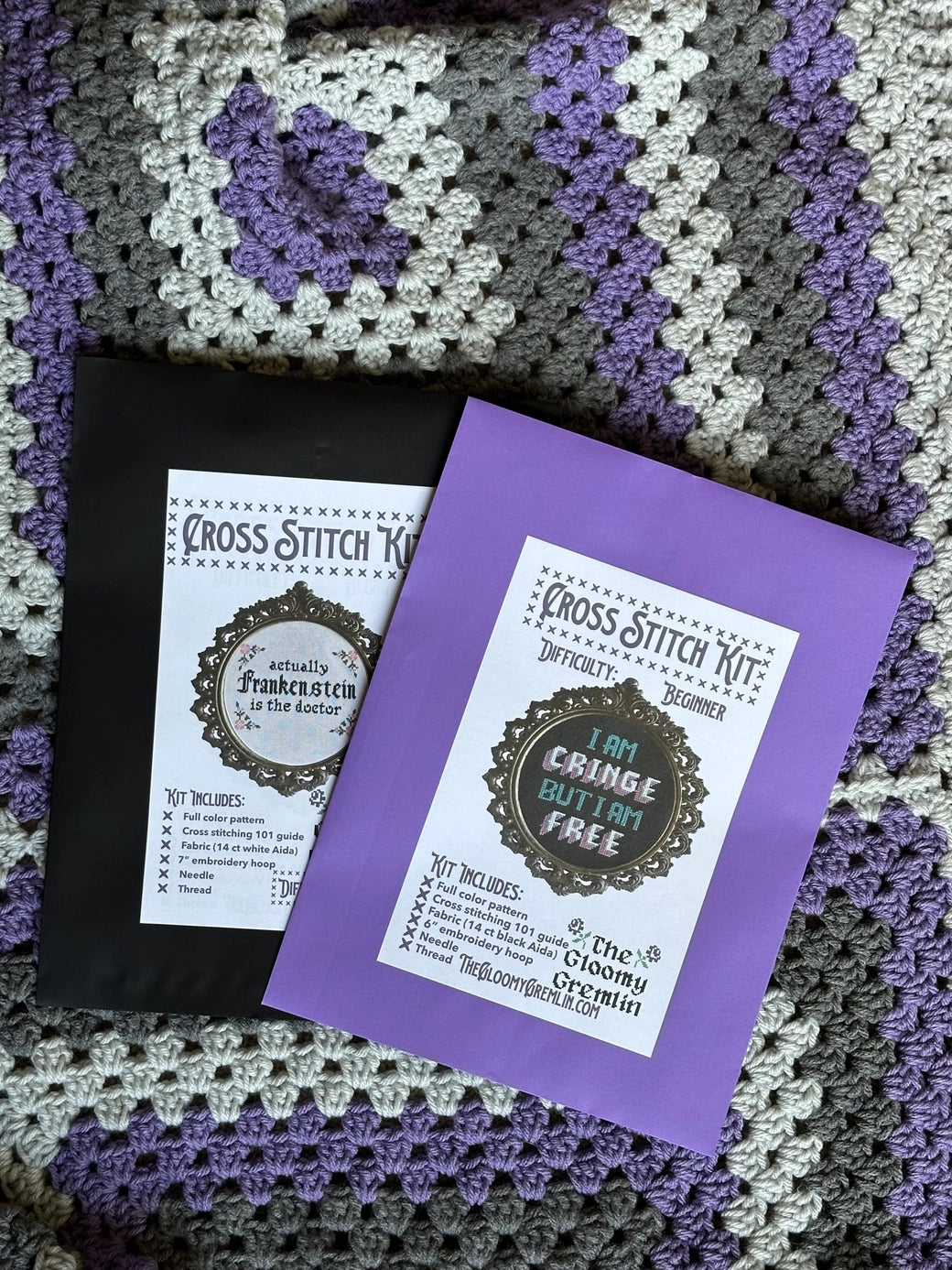 Two cross stitch kits on a crocheted blanket with purple and gray patterns.