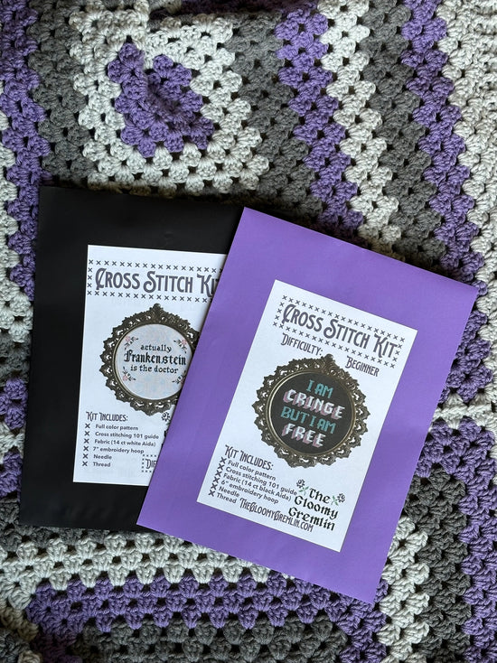 Two cross stitch kits on a crocheted blanket with purple and gray patterns.
